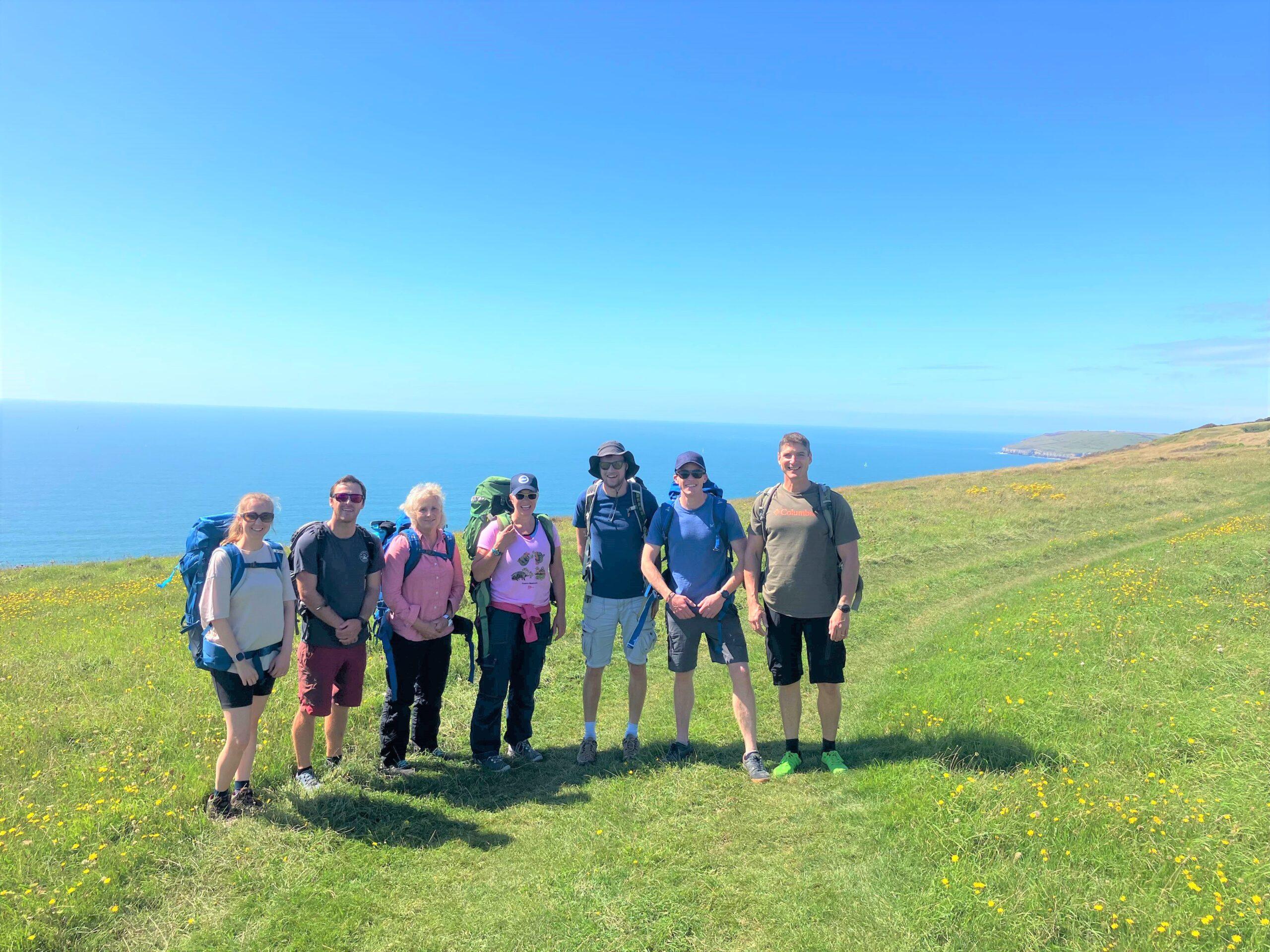 A group of 7 people are stood at the top of a grassy cliff looking over the sea.